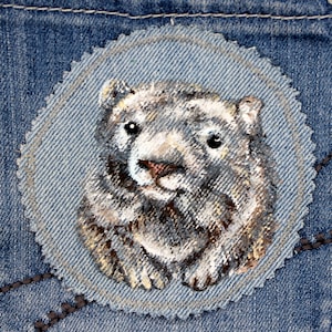 Könnte beinhalten: Nahaufnahme eines handgemalten Jeans-Patches mit einem grauen und braunen Wombat mit weißer Nase und schwarzen Augen. Der Patch ist rund und hat einen gewellten Rand.