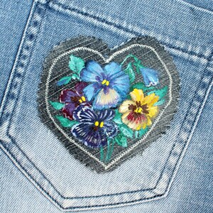 May include: A heart-shaped patch with a painted design of purple, blue, and yellow pansies on a denim background.