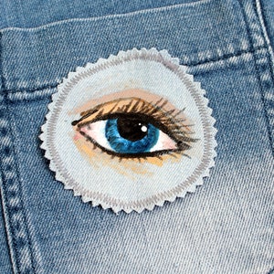 May include: A blue eye with brown eyelashes and a light brown eyebrow is painted on a white fabric circle patch. The patch is sewn onto a blue denim background.