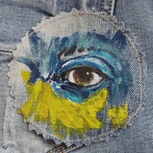 Peut inclure: Gros plan sur un œil bleu peint sur un tissu en jean. L'œil est entouré de peinture jaune et bleue, créant un motif floral.