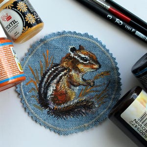 May include: A circular denim patch featuring a hand-painted chipmunk surrounded by tall grass. The patch has a scalloped, stitched edge and is surrounded by various bottles of fabric paint and two paintbrushes on a white surface.