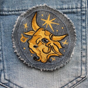 Könnte beinhalten: Ein blauer Jeans-Patch mit einem gestickten gelben Stierkopf und einem Sternmuster.