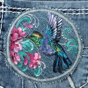 Pode incluir: Um remendo de brim azul com um beija-flor pintado à mão em voo, rodeado de flores cor-de-rosa e folhagem verde.