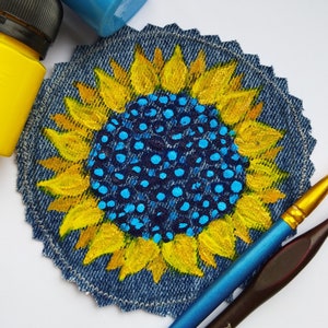 Peut inclure: Un patch en jean peint à la main représentant un tournesol avec un centre bleu et jaune. Le tournesol est peint sur un fond en jean bleu.