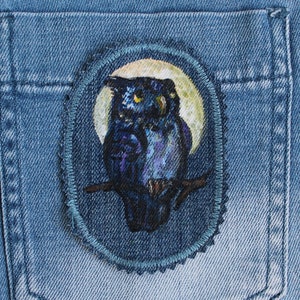 Könnte beinhalten: Eine blaue Jeans-Tasche mit einem runden gestickten Aufnäher, der einen schwarzen Eulen zeigt, der auf einem Ast vor einem gelben Mond sitzt.