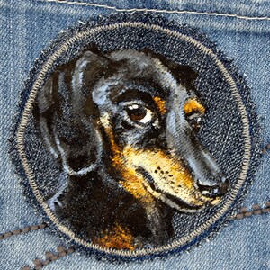 Puede incluir: Un perro dachshund negro y marrón pintado en tela vaquera. La cabeza del perro está de perfil, mirando hacia la derecha. Los ojos del perro son marrones y la nariz es negra. El perro está rodeado de un óvalo cosido.