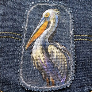 May include: A denim patch featuring a detailed illustration of a pelican. The bird is depicted with a long orange beak, gray and white feathers, and a yellow eye. The patch is stitched onto a blue denim background.