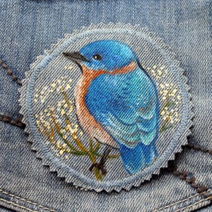 Op de afbeelding: Een ronde denim patch met een gedetailleerde schildering van een blauwe vogel die op een tak zit met kleine witte bloemen. De vogel heeft blauwe en oranje veren, een zwarte snavel en een klein oog. De patch is op een denim achtergrond genaaid.