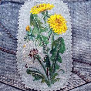 Op de afbeelding: Een denim patch met een schildering van paardenbloemen. De patch toont heldergele paardenbloem bloemen, groene bladeren en een witte paardenbloem zaadhoofd. De ovaalvormige patch heeft een lichtblauwe rand en is op een denim zak genaaid.