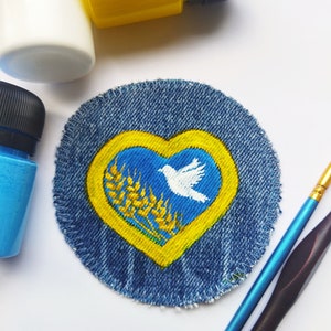 Peut inclure: Un patch en jean bleu avec un motif en forme de cœur jaune. Le cœur contient un fond bleu avec une colombe blanche et des épis de blé.