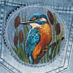 Könnte beinhalten: Ein handgemalter Jeans-Patch mit einem blauen und orangefarbenen Eisvogel, der auf einem Ast mit grünen Schilf sitzt. Der Patch ist von einem ausgefransten Rand umgeben.