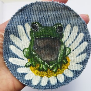Op de afbeelding: Een groene kikker met bruine ogen zit op een witte madeliefje geverfd op een blauwe denim patch. De madeliefje heeft een gele kern.
