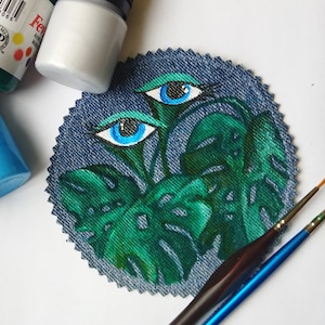 Puede incluir: Un parche circular de mezclilla con un diseño pintado de hojas verdes de monstera y dos ojos azules. El parche está rodeado por una puntada decorativa.