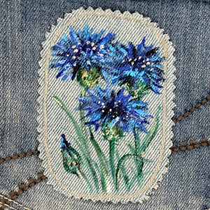 Pode incluir: Um close-up de um remendo de ganga com um desenho pintado à mão de açafrão azul. As flores são pintadas em tons de azul e verde, com acentos brancos. O remendo é costurado em um fundo de ganga.