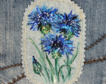 May include: A close-up of a denim patch with a hand-painted design of blue cornflowers. The flowers are painted in shades of blue and green, with white accents. The patch is sewn onto a denim background.