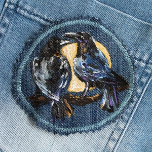 May include: A denim patch with an embroidered design of two black crows perched on a branch. The crows are facing each other, with one looking to the left and the other looking to the right.