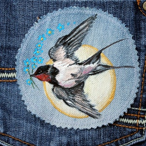 May include: A blue denim patch with a hand-painted image of a bird with a yellow beak and blue flowers. The bird is flying with its wings spread and has a white breast.