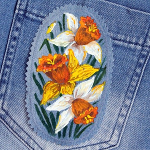 May include: A hand-painted denim patch with three yellow and white daffodils with orange centers. The patch is on a blue denim background.