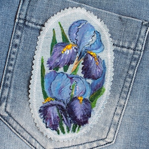 May include: A blue denim pocket with a white embroidered patch featuring a cluster of purple and blue irises with green leaves.