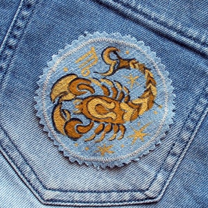 Pode incluir: Um remendo de ganga azul com um design de escorpião bordado em branco. O escorpião é contornado em branco e preenchido com amarelo e marrom. O fundo é azul claro com estrelas brancas.