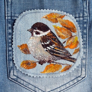 May include: A hand-painted denim patch depicting a sparrow perched on a branch with autumn leaves. The sparrow is brown, grey, and white with black stripes. The leaves are orange and yellow.