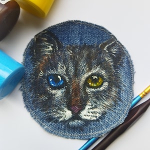 Peut inclure: Un portrait de chat peint à la main sur un patch en jean. Le chat a les yeux bleus et jaunes et un nez brun. Le patch est entouré de pinceaux et de pots de peinture.
