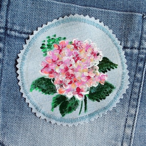 Könnte beinhalten: Nahaufnahme eines Jeans-Patches mit einem handgemalten Design einer rosa und weißen Hortensie mit grünen Blättern.