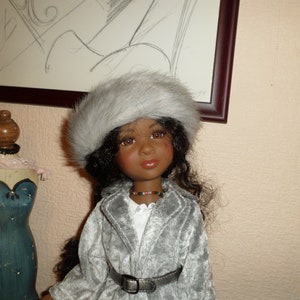 Peut inclure: Poupée à la peau foncée, portant un chapeau de fourrure gris et un manteau assorti avec une ceinture argentée. La poupée a de longs cheveux bouclés foncés et porte un collier de perles. Un mannequin décoratif et une esquisse encadrée sont en arrière-plan.