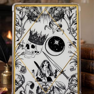 Puede incluir: Funda transparente para teléfono con borde dorado e ilustración en blanco y negro. El diseño presenta una calavera con corona, una luna creciente con estrella, una mujer escribiendo, flores y una espada. El conjunto está enmarcado por motivos florales.