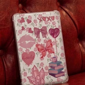 Könnte beinhalten: Transparente Handyhülle mit weißem Hintergrund, verziert mit rosa Herzen, Blumen, einem Schmetterling, Lippen, einer Schleife und dem Text "XOXO". Die Hülle hat auch einen Stapel Bücher, eine Tasse und Kristalle.