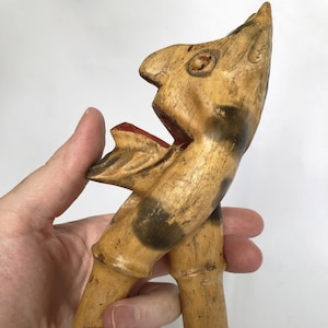 Op de afbeelding: Een houten notenkraker gebeeldhouwd in de vorm van een vogel met een zwart en bruin gevlekt patroon. De bek van de vogel is open en rood geverfd. De notenkraker wordt in een hand gehouden.