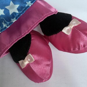 May include: Close-up of a doll's legs and feet. The doll's feet are covered in shiny, pink satin slippers with small, light pink bows. The doll's legs are covered in a blue fabric with white stars.