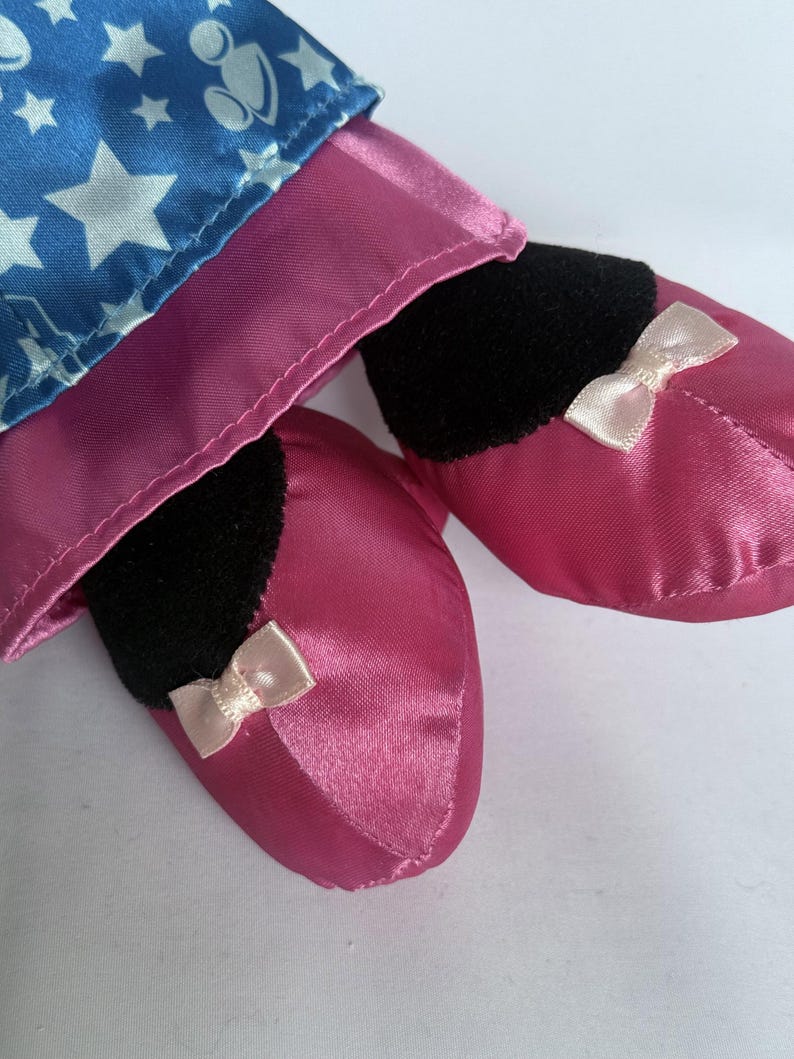 May include: Close-up of a doll's legs and feet. The doll's feet are covered in shiny, pink satin slippers with small, light pink bows. The doll's legs are covered in a blue fabric with white stars.
