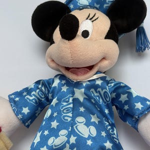 May include: A plush Minnie Mouse doll wearing a blue graduation gown and cap. The gown and cap are decorated with white stars and the year "2014". The doll has black ears, a pink face, and a smiling expression.