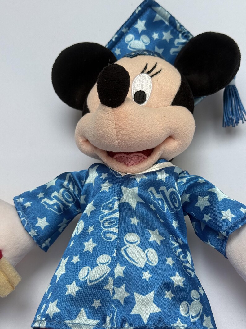 May include: A plush Minnie Mouse doll wearing a blue graduation gown and cap. The gown and cap are decorated with white stars and the year "2014". The doll has black ears, a pink face, and a smiling expression.
