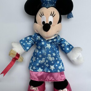 May include: A plush Minnie Mouse doll wearing a blue graduation cap and gown with white stars and Mickey Mouse silhouettes. The gown has the year "2014" printed on it. She holds a rolled diploma with a red ribbon and wears pink shoes.
