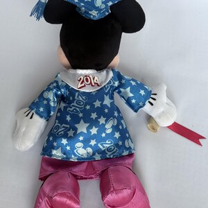 May include: A plush Minnie Mouse doll wearing a blue graduation gown with white stars and the year "2014" on the collar. The doll has pink shoes, white gloves, and a matching graduation cap. A rolled diploma with a red ribbon is in her hand.