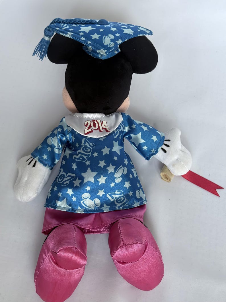 May include: A plush Minnie Mouse doll wearing a blue graduation gown with white stars and the year "2014" on the collar. The doll has pink shoes, white gloves, and a matching graduation cap. A rolled diploma with a red ribbon is in her hand.