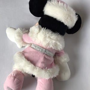 May include: Pink plush Minnie Mouse wearing a white fur trimmed coat and hat. The plush toy has a silver glitter belt and a white fur trim around the cuffs of the sleeves.