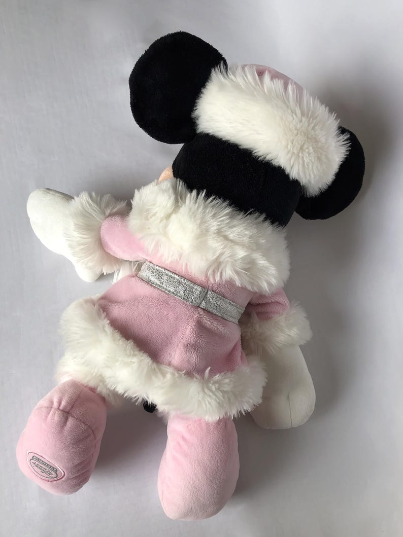 May include: Pink plush Minnie Mouse wearing a white fur trimmed coat and hat. The plush toy has a silver glitter belt and a white fur trim around the cuffs of the sleeves.