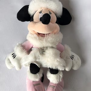 May include: A plush toy of Minnie Mouse wearing a white winter coat and hat with pink boots.