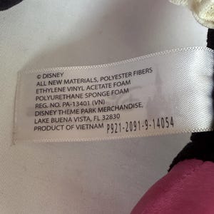 May include: A white tag with black text, featuring the Disney logo and product information. The tag lists materials like polyester fibers and foam, along with a product code and the location of Disney Theme Park Merchandise in Lake Buena Vista, Florida.