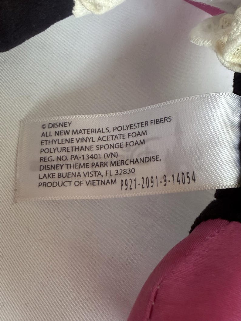 May include: A white tag with black text, featuring the Disney logo and product information. The tag lists materials like polyester fibers and foam, along with a product code and the location of Disney Theme Park Merchandise in Lake Buena Vista, Florida.
