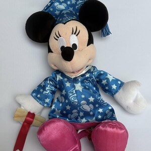 May include: A Minnie Mouse plush doll wearing a blue graduation cap and gown with white stars and the year "2014" printed on the gown. The doll has black ears, a pink nose, and pink satin shoes. It holds a rolled-up diploma with a red ribbon.