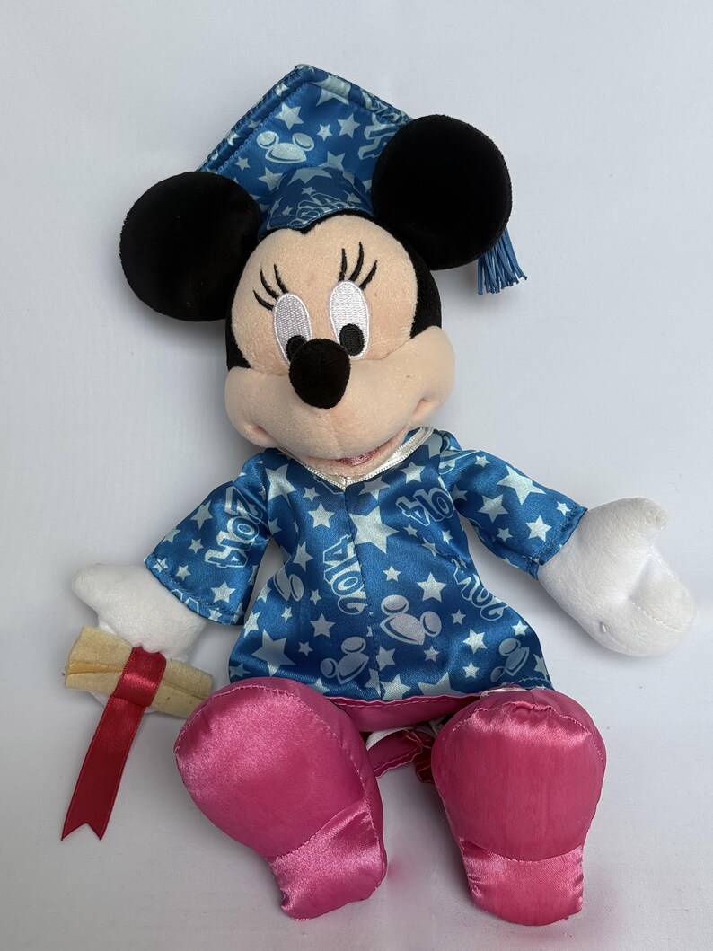 May include: A Minnie Mouse plush doll wearing a blue graduation cap and gown with white stars and the year "2014" printed on the gown. The doll has black ears, a pink nose, and pink satin shoes. It holds a rolled-up diploma with a red ribbon.