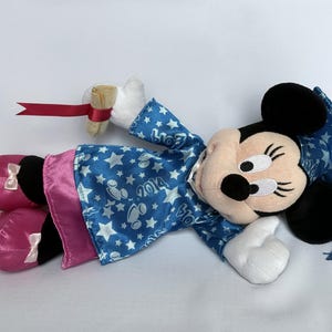 May include: A Minnie Mouse plush doll wearing a blue graduation gown with white stars and the year 2014. The doll has pink satin shoes with white bows and is holding a diploma with a red ribbon.