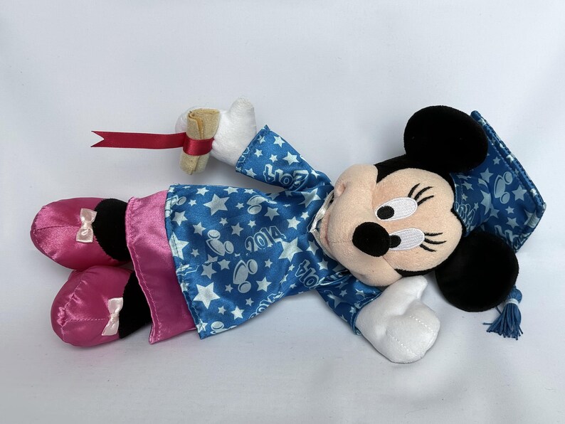 May include: A Minnie Mouse plush doll wearing a blue graduation gown with white stars and the year 2014. The doll has pink satin shoes with white bows and is holding a diploma with a red ribbon.