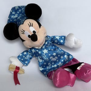 May include: A Minnie Mouse plush doll wearing a blue pajama set with white stars and Mickey Mouse silhouettes. The doll has black ears, a pink skirt, and pink shoes. It holds a scroll with a red ribbon.