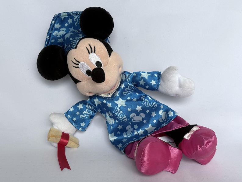 May include: A Minnie Mouse plush doll wearing a blue pajama set with white stars and Mickey Mouse silhouettes. The doll has black ears, a pink skirt, and pink shoes. It holds a scroll with a red ribbon.
