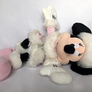 May include: A plush toy of Minnie Mouse dressed in a white and pink winter outfit with a white fur trim. She is wearing pink boots and has a black nose and eyes.